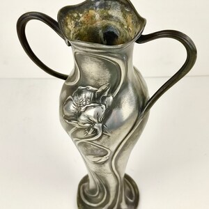 K&ouml;nnte beinhalten: Eine silberne Zinnvase mit zwei Henkeln und einem floralen Art-Nouveau-Design. Die Vase ist mit einer stilisierten Blume und Bl&auml;ttern verziert.
