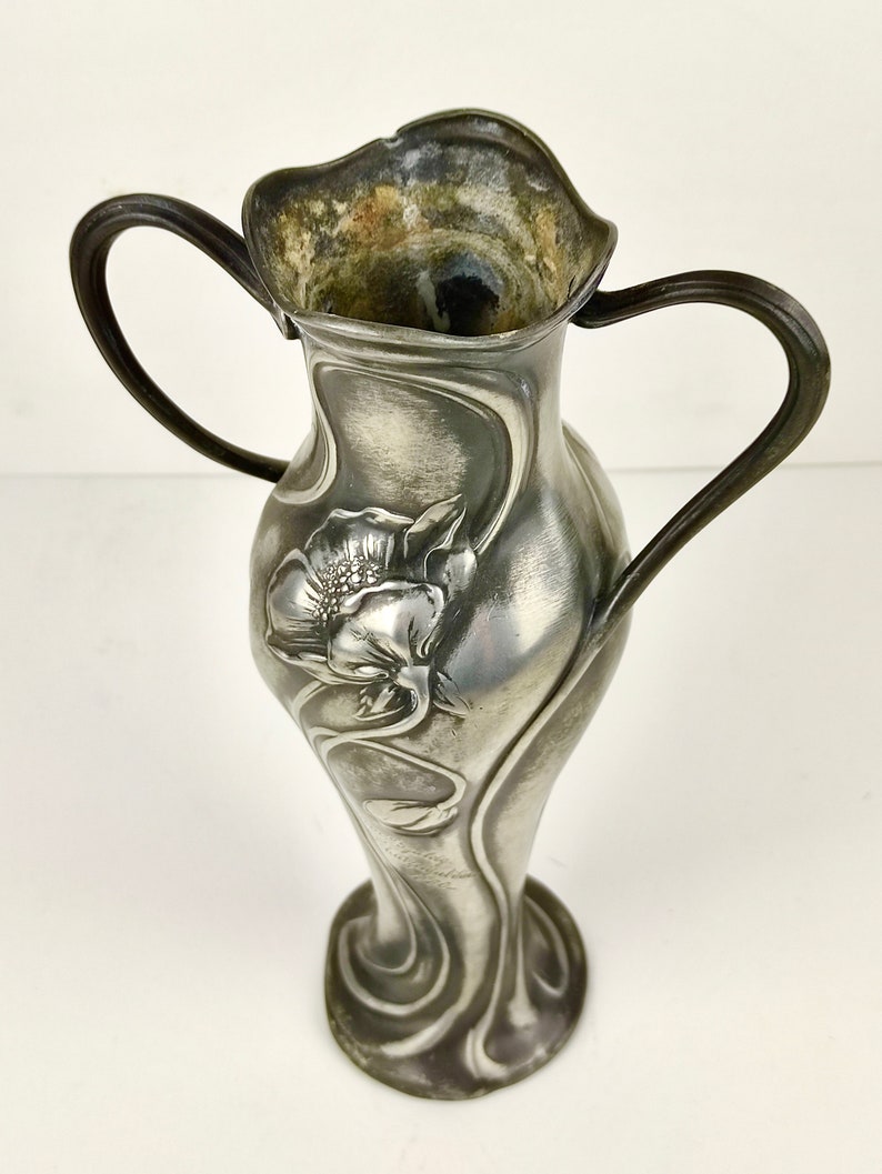 K&ouml;nnte beinhalten: Eine silberne Zinnvase mit zwei Henkeln und einem floralen Art-Nouveau-Design. Die Vase ist mit einer stilisierten Blume und Bl&auml;ttern verziert.