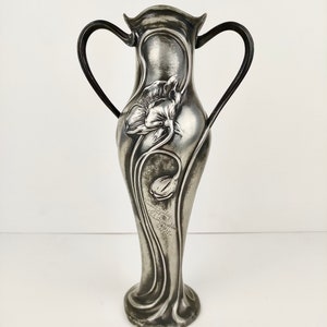 K&ouml;nnte beinhalten: Eine silberne Zinnvase mit zwei Griffen und einem Blumendesign. Die Vase ist mit einer stilisierten Blume und der Inschrift "Atelier  Schmid  M&uuml;nchen  1902" verziert.