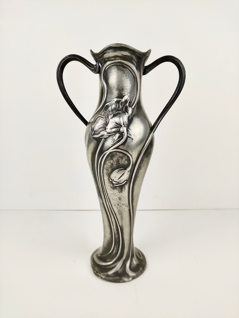 K&ouml;nnte beinhalten: Eine silberne Zinnvase mit zwei Griffen und einem Blumendesign. Die Vase ist mit einer stilisierten Blume und der Inschrift "Atelier  Schmid  M&uuml;nchen  1902" verziert.