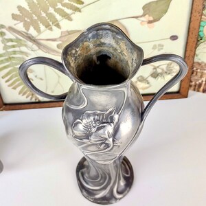 K&ouml;nnte beinhalten: Eine silberne Vase im Jugendstil mit floralem Design. Die Vase hat zwei Henkel und eine breite &Ouml;ffnung.