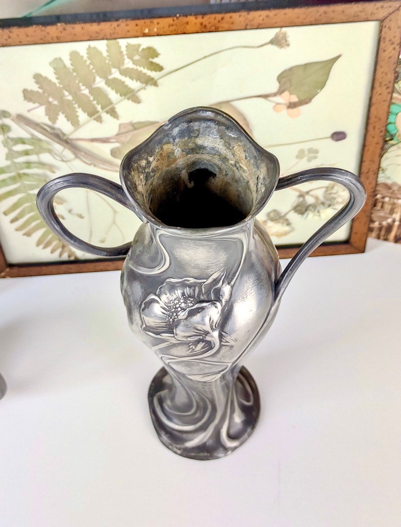 K&ouml;nnte beinhalten: Eine silberne Vase im Jugendstil mit floralem Design. Die Vase hat zwei Henkel und eine breite &Ouml;ffnung.