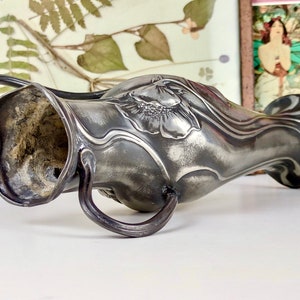 K&ouml;nnte beinhalten: Eine silberne Zinnvase mit einem Jugendstil-Design, das eine stilisierte Blume zeigt. Die Vase hat zwei Henkel und einen leicht geschwungenen K&ouml;rper.