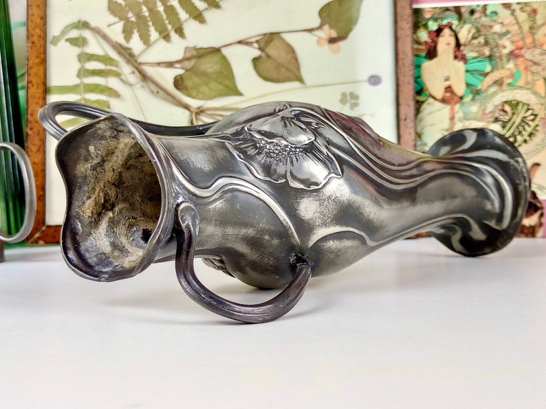 K&ouml;nnte beinhalten: Eine silberne Zinnvase mit einem Jugendstil-Design, das eine stilisierte Blume zeigt. Die Vase hat zwei Henkel und einen leicht geschwungenen K&ouml;rper.