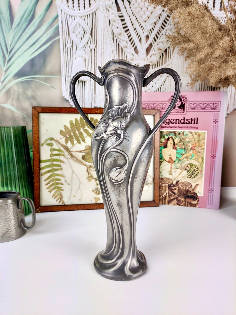 K&ouml;nnte beinhalten: Eine silberne Vase im Jugendstil mit zwei Henkeln und einem floralen Design. Die Vase ist mit einer stilisierten Blume und Bl&auml;ttern verziert.