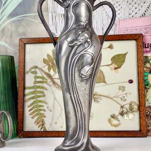 K&ouml;nnte beinhalten: Eine silberne Zinnvase mit floralem Design und zwei Henkeln. Die Vase ist mit einer stilisierten Blume verziert und tr&auml;gt die Inschrift "Emile Gall&eacute;, Nancy, 1900".
