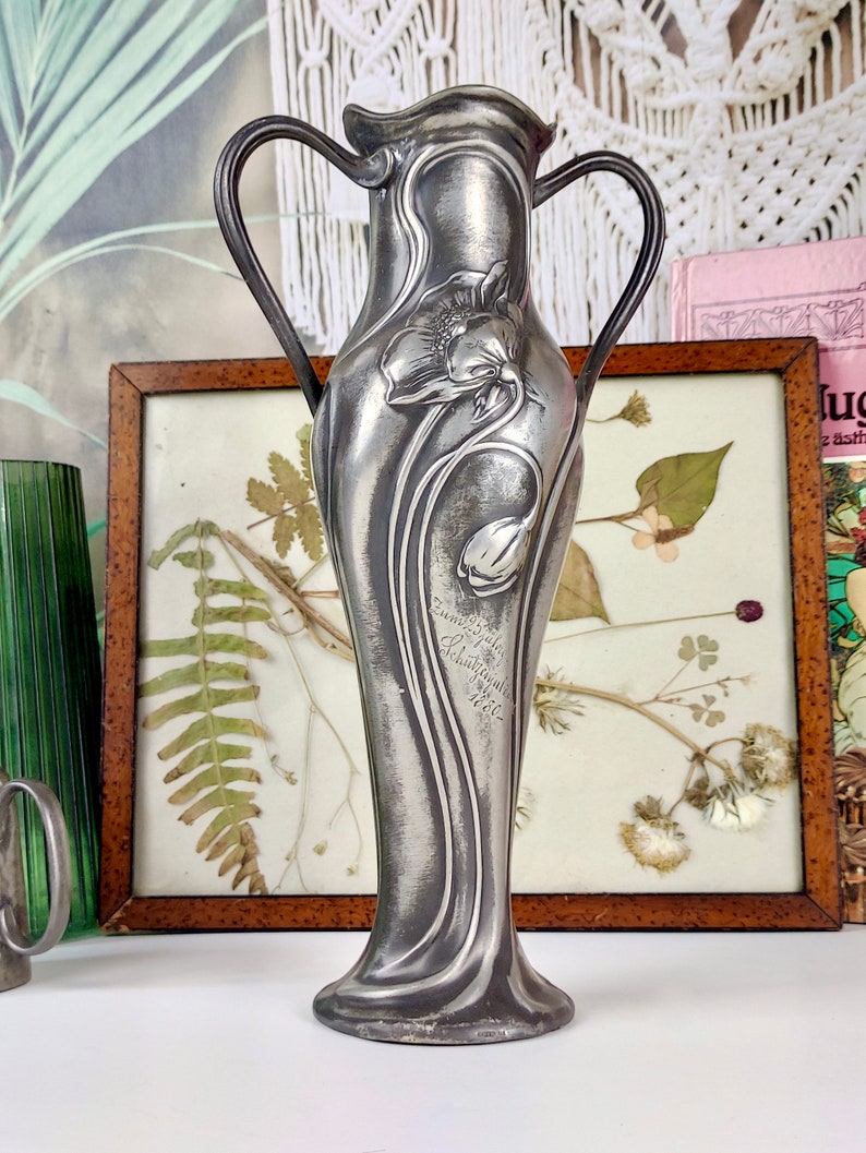 K&ouml;nnte beinhalten: Eine silberne Zinnvase mit floralem Design und zwei Henkeln. Die Vase ist mit einer stilisierten Blume verziert und tr&auml;gt die Inschrift "Emile Gall&eacute;, Nancy, 1900".