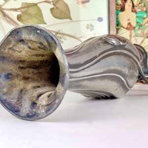 K&ouml;nnte beinhalten: Eine silberne Zinnvase mit einem strukturierten, wellenf&ouml;rmigen Design und einer breiten, ausgestellten &Ouml;ffnung. Die Vase hat einen Henkel, der wie eine Schlaufe geformt ist.