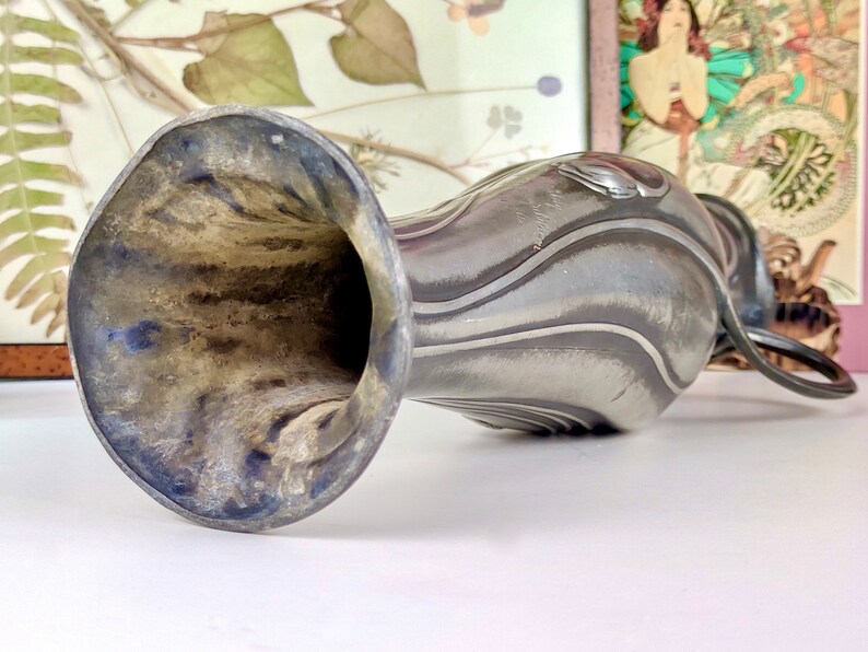 K&ouml;nnte beinhalten: Eine silberne Zinnvase mit einem strukturierten, wellenf&ouml;rmigen Design und einer breiten, ausgestellten &Ouml;ffnung. Die Vase hat einen Henkel, der wie eine Schlaufe geformt ist.