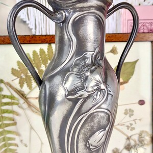 K&ouml;nnte beinhalten: Eine silberne Zinnvase mit zwei Henkeln, verziert mit einem gepr&auml;gten Blumendesign aus einer Mohnblume und einer Tulpe. Die Vase ist signiert "Zum 25 j&auml;hrigen Schuljubil&auml;um 1912".