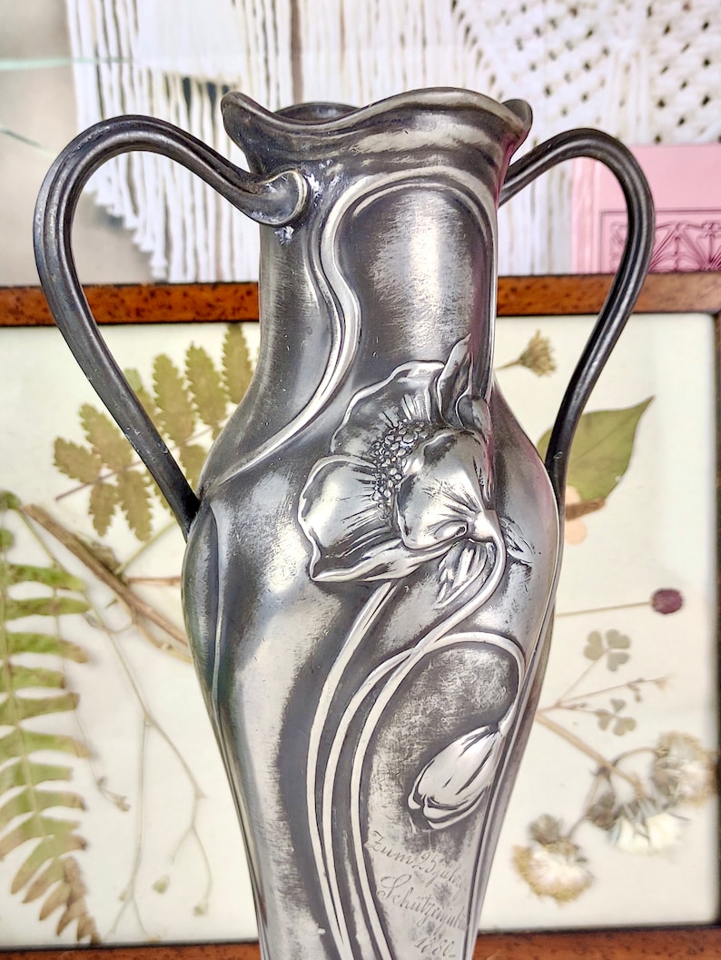 K&ouml;nnte beinhalten: Eine silberne Zinnvase mit zwei Henkeln, verziert mit einem gepr&auml;gten Blumendesign aus einer Mohnblume und einer Tulpe. Die Vase ist signiert "Zum 25 j&auml;hrigen Schuljubil&auml;um 1912".