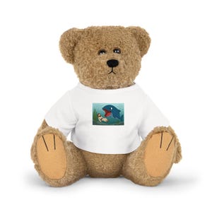 Puede incluir: Un oso de peluche marrón con una camiseta blanca con un dibujo animado. El dibujo representa a una persona en el agua con un tiburón azul. El oso tiene una nariz negra y patas marrones.
