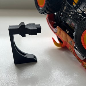 May include: A black 3D-printed stand designed to hold a toy monster truck. The stand is a geometric shape with a curved support and a flat base. The monster truck is red, orange, and yellow with large black tires.
