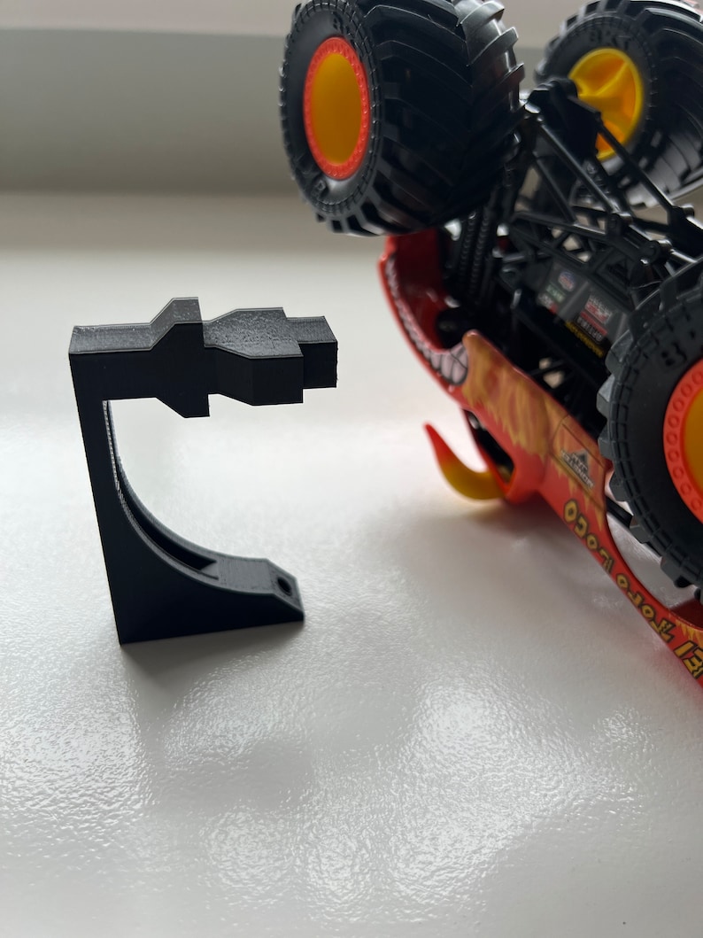 May include: A black 3D-printed stand designed to hold a toy monster truck. The stand is a geometric shape with a curved support and a flat base. The monster truck is red, orange, and yellow with large black tires.