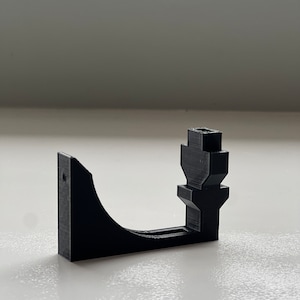 May include: A black 3D-printed bracket with a curved arm and a vertical post. The bracket has a rectangular base with a hole for mounting. The design is simple and functional, likely for holding or supporting an object.