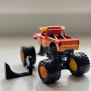 May include: A red and orange toy monster truck with the words "El Toro Loco" printed on the side. The truck has large black tires with yellow and orange accents. A black plastic piece is next to the truck.