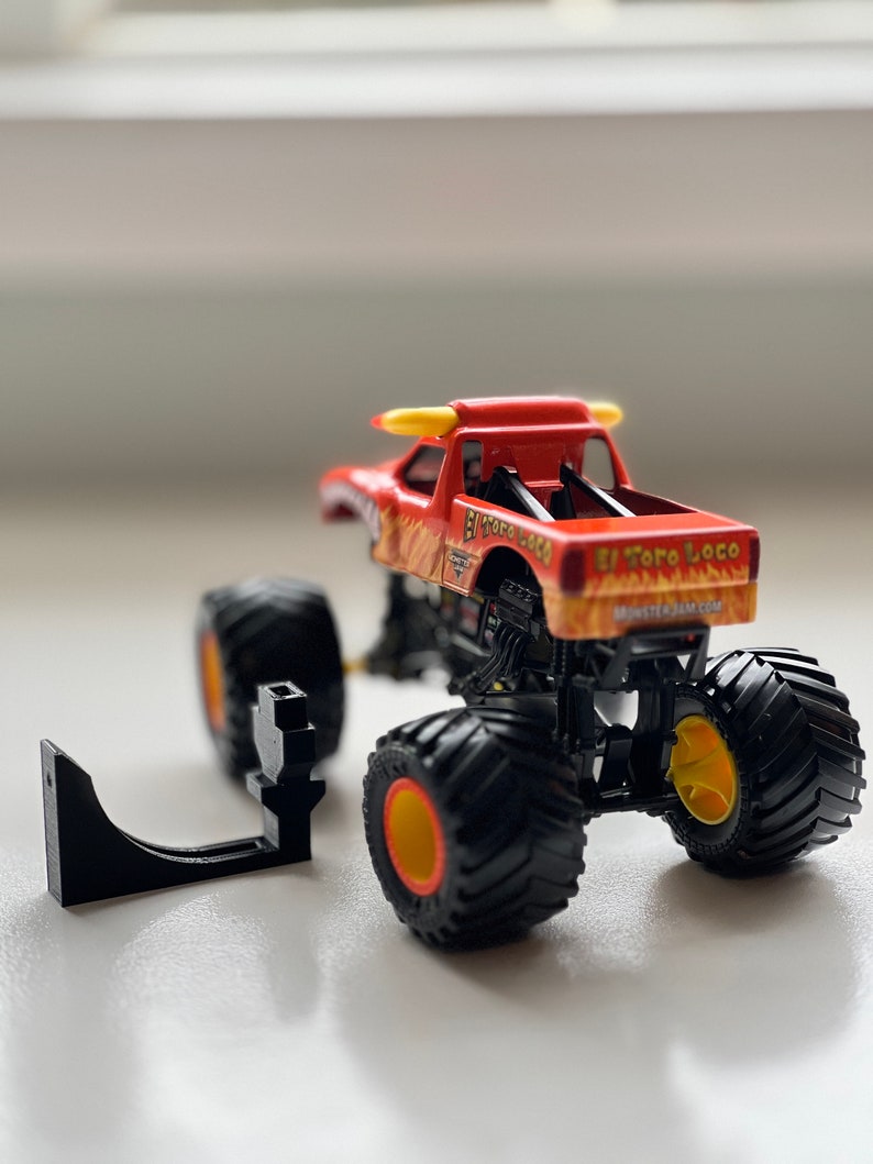 May include: A red and orange toy monster truck with the words "El Toro Loco" printed on the side. The truck has large black tires with yellow and orange accents. A black plastic piece is next to the truck.