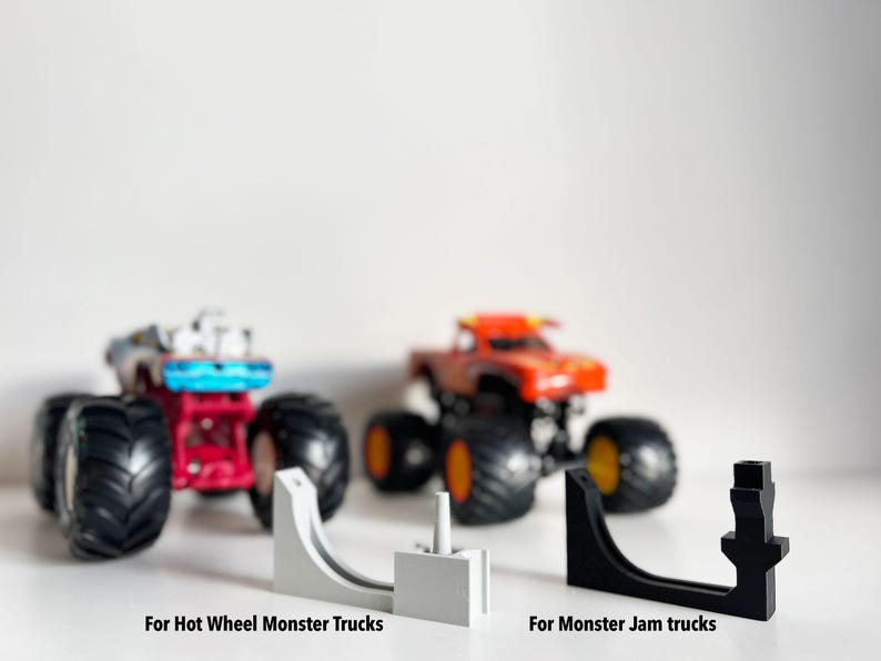 May include: Image features two toy monster trucks, one red and one blue, with large black tires. In front are two ramps, one white and one black, labeled "For Hot Wheel Monster Trucks" and "For Monster Jam trucks."