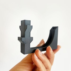 May include: A dark gray, 3D-printed bracket with a unique architectural design. The bracket features a series of stacked, geometric shapes and a curved support arm. The object is held in a hand against a white background.
