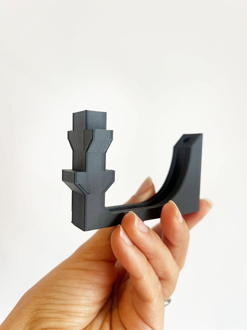 May include: A dark gray, 3D-printed bracket with a unique architectural design. The bracket features a series of stacked, geometric shapes and a curved support arm. The object is held in a hand against a white background.