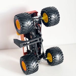 May include: A red and black monster truck toy with large black tires and yellow and orange accents. The toy truck is upside down, showcasing its undercarriage and wheel design. The tires have a deep tread pattern.