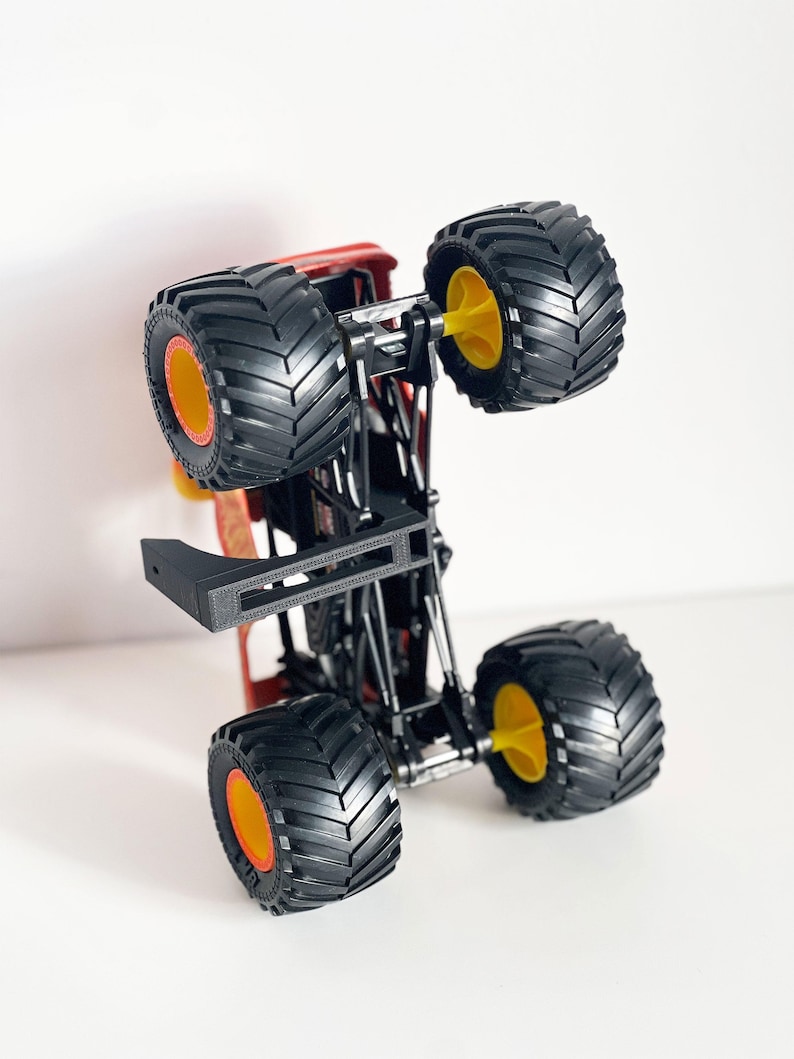 May include: A red and black monster truck toy with large black tires and yellow and orange accents. The toy truck is upside down, showcasing its undercarriage and wheel design. The tires have a deep tread pattern.