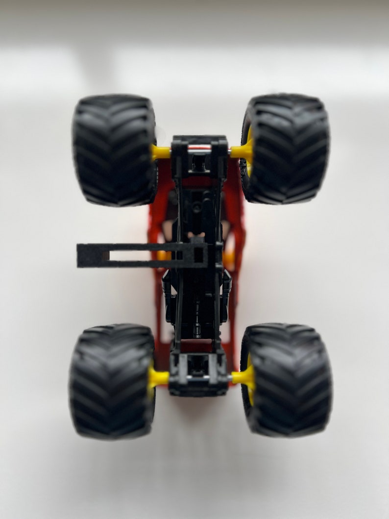 May include: A red and black toy monster truck with four large black tires and yellow wheel hubs. The truck's chassis is visible, showcasing its mechanical details. The tires have a deep tread pattern.