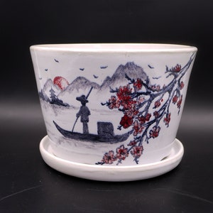 Puede incluir: Maceta de cerámica blanca con una escena pintada a mano de una persona en un barco con montañas y un sol rojo en el fondo. La maceta tiene un platillo a juego.