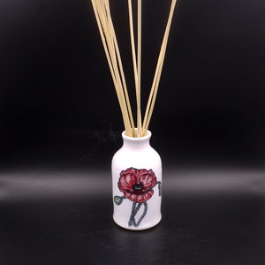 Op de afbeelding: Witte keramische diffuserfles met een rood klaproosontwerp en houten diffuserstokjes.