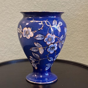 May include: A cobalt blue vase featuring a hand-painted floral pattern with light blue petals, dark blue centres, and green leaves. The design is accented with metallic gold outlines on the flowers, leaves, and branches, with gold trim on the rim and base.