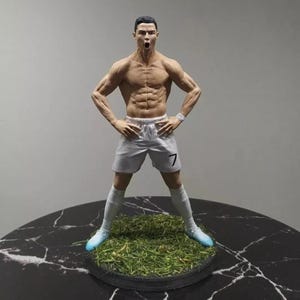 Puede incluir: Figura detallada de un futbolista sin camiseta, con pantalones cortos blancos con el número 7, calcetines blancos y zapatillas azules. La figura está en una pose dinámica, sobre una base circular con césped artificial.