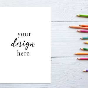 以下が含まれることがあります： 白い紙に「your design here」という文字が書かれています。紙は白い木製の表面に置かれています。画像の右側には、いくつかの色鉛筆が縦に並んでいます。