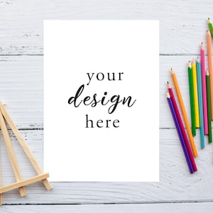 以下が含まれることがあります： 白い木製の表面に「your design here」というテキストが書かれた白い紙。小さな木製イーゼルと色鉛筆の束が表面にある。