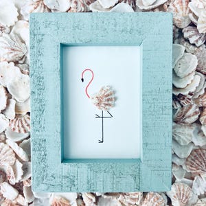 Könnte beinhalten: Ein rechteckiger hellblauer Holzrahmen mit weißem Hintergrund und einer minimalistischen Flamingo-Illustration. Der Körper des Flamingos besteht aus einer strukturierten Muschel, während Hals, Kopf und Beine mit dünnen Linien gezeichnet sind.