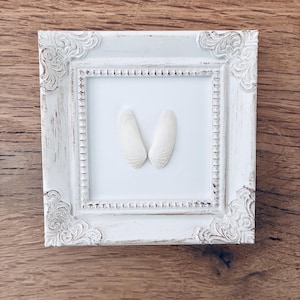 May include: A square, white, ornate picture frame with a distressed finish. Inside the frame, two white seashells are arranged on a white background. The frame sits on a wooden surface.