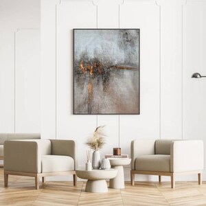 May include: Two beige armchairs with wooden legs and a white coffee table with a vase of dried pampas grass and a book on top. A framed abstract painting with gray and brown tones hangs on the wall behind the coffee table.