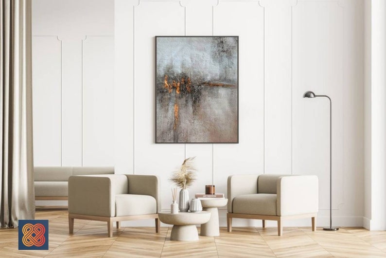 May include: Two beige armchairs with wooden legs and a white coffee table with a vase of dried pampas grass and a book on top. A framed abstract painting with gray and brown tones hangs on the wall behind the coffee table.