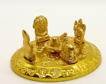 Könnte beinhalten: Ein goldfarbener Metallständer mit runder Basis und vier kleinen Figuren darauf. Die Figuren stellen hinduistische Gottheiten dar, darunter Shiva, Vishnu und Brahma. Die Basis ist mit Sanskrit-Text beschriftet.