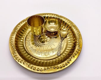 May include: A gold-colored brass plate with a raised rim and a decorative pattern. The plate has three compartments, one for a small cup, one for a small bowl, and one for a spoon. The plate also has a sunburst design with the word "Om" in the center. The plate is inscribed with the text "Shanti,  Vishva Shanti,  Sarva Shanti".