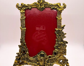 May include: A gold-toned metal picture frame with an ornate design. The frame has a red velvet backing and stands on three legs.