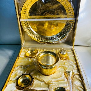 May include: A gold-colored metal set of ceremonial items in a gold-colored box with a satin lining. The set includes a large plate, a bowl, a small cup, a spoon, a small pot, and a small incense burner.