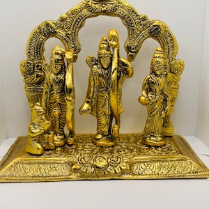 May include: A gold-colored metal statue of three Hindu deities, Rama, Sita, and Lakshmana, standing on a decorative base. The statue is intricately detailed and features a traditional archway.