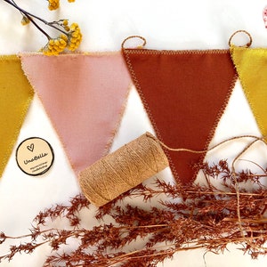 May include: A bunting banner with four triangular flags in shades of yellow, pink, brown, and mustard yellow. The flags are sewn together with a brown twine. The bunting is on a white background with a small wooden sign that says "UnaBella" with a heart symbol.