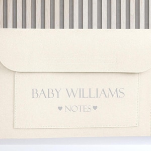 May include: A cream-coloured felt document folder with a flap closure. The front features the text BABY WILLIAMS centred above the word NOTES, which is flanked by two small heart icons. The material has a soft, textured appearance.