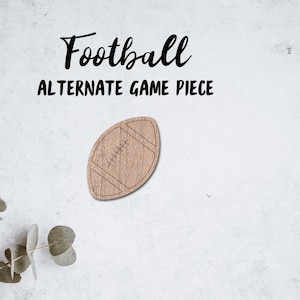 以下が含まれることがあります： 縫い目のディテールが施されたフットボールの形をした木製フットボールゲームピース。ピースは、その上に「Football Alternate Game Piece」というテキストが書かれた白い背景の上にあります。