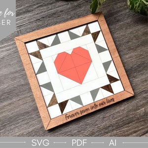 May include: A wooden framed square with a white background and a red heart in the center. The frame is made of wood with a brown stain. The background is white with a pattern of gray and brown triangles. The text "forever sewn into our lives" is written below the heart.