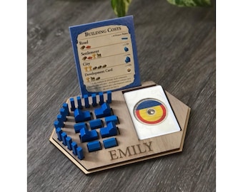 May include: Wooden game organizer with blue game pieces and a card. The organizer has a slot for cards and a section for game pieces. The name "EMILY" is engraved on the front. A card with building costs is displayed.