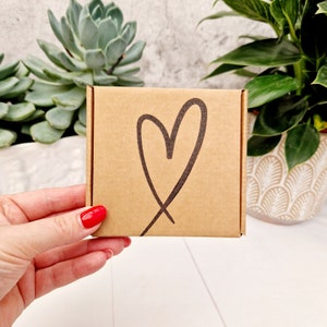 May include: A small brown cardboard box with a hand-drawn heart design. The box is being held by a hand with red nail polish.