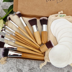 Op de afbeelding: Een set van 10 bamboe make-up kwasten met zilveren metalen ferrules en zwarte haren. De kwasten zijn gerangschikt in een doos met een set ronde katoenen make-up remover pads in een gaas zak.