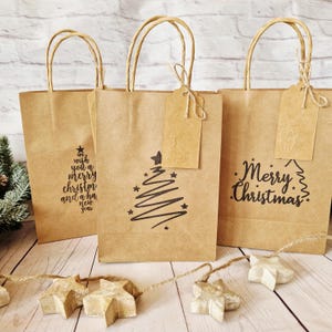Peut inclure: Trois sacs cadeaux en papier brun avec des poignées torsadées. Chaque sac présente un motif de Noël noir différent : un sapin de Noël, les mots "Merry Christmas" et le texte "We wish you a Merry Christmas and a Happy New Year".